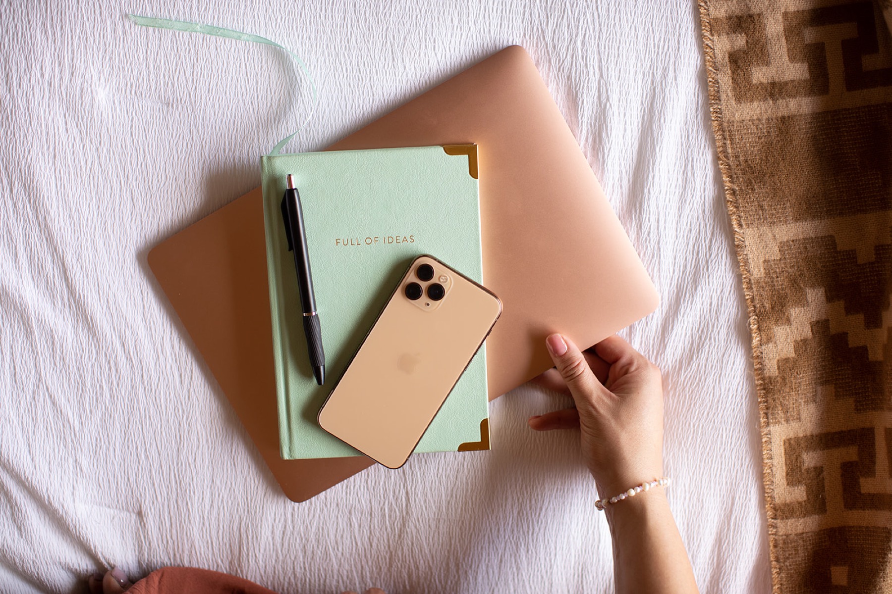 An overhead flatlay of a sage-green journal and rose-gold phone on soft bed linens, with a hand resting alongside